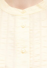 Chemise de couleur crème avec des rayures verticales texturées et trois boutons blancs sur le devant, présentant un col arrondi.