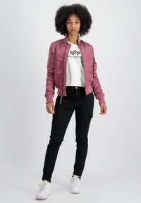 Veste bomber rose avec des poignets côtelés, des poches zippées et un patch logo, associée à un pantalon cargo noir et des baskets blanches.