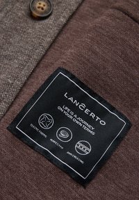 Brązowy materiał o tkanej strukturze z czarną metką informacyjną. Metka zawiera ikony: elastyczny materiał, idealne dopasowanie, przeciw zagnieceniom. Widoczny detal guzika.