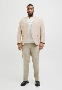 Blazer en lin beige avec un pantalon assorti, porté sur un polo blanc. Texture lisse, coupe à un bouton et poches latérales. Chaussures noires.
