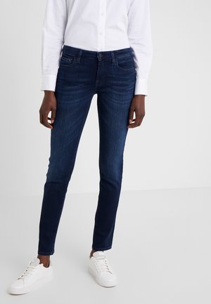 Person iført mørkeblå slim-fit jeans, hvid skjorte med knapper og hvide sneakers, stående mod en ensartet lysegrå baggrund.