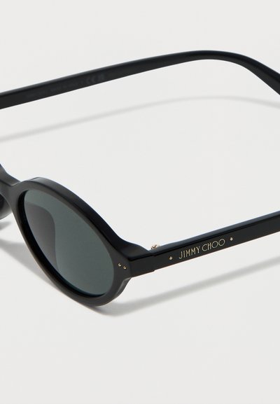 Μαύρα γυαλιά ηλίου με στρογγυλό πλαίσιο και σκοτεινούς φακούς, με το χρυσό λογότυπο "JIMMY CHOO" στο βραχίονα του πλαισίου, σε λευκό φόντο.