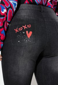 Vaqueros de mezclilla negra con un diseño de bolsillo trasero que incluye detalles pintados en rojo, como "xoxo" y un corazón, sobre una tela texturizada.