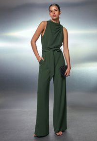 Grön jumpsuit med draperad halsdesign, vida byxor, midjebälte och sidfickor, accessoarer med en liten svart kuvertväska.
