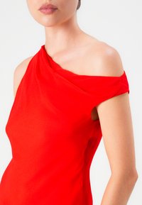 Mujer vestida con un vestido rojo de un solo hombro, mostrando el hombro y el brazo desnudos contra un fondo blanco liso.