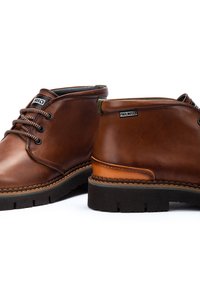 Bottines en cuir marron avec un bout arrondi, des accents contrastants, des lacets ronds et une semelle en caoutchouc texturée. Étiquette de marque visible.