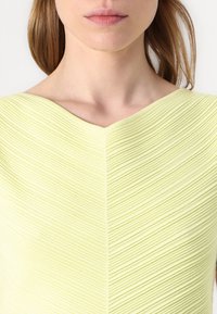 Lys gul topp med V-hals, med teksturerte ribbestrikkede detaljer i et chevron-mønster. Smidig stoff og tilpasset design er synlig.