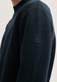 Pull en laine bleu marine avec un tissu texturé, un col côtelé et un petit logo sur la manche. Coupe ajustée, design minimaliste.