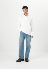Vit zip-up sweatshirt med en grön logo, kombinerad med ljusblå vida jeans och svarta sneakers. Minimalistisk design, slät textur.