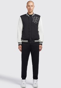 Zwarte bomberjack met witte mouwen, gestreepte manchetten en een geborduurde letter. Combinatie met donkere broek en witte sneakers.