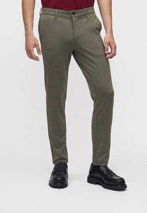 Homme portant un pantalon slim vert olive, des mocassins en cuir noirs et une chemise rouge, debout devant un fond blanc uni.