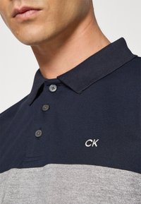 Navy og grå stripete polo skjorte med krage, med tre knapper og brodert "CK"-logo på venstre bryst. Teksturert stoff.