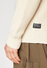 Jersey de punto en color blanco roto, con una textura acanalada y puños. Incluye una etiqueta negra con el nombre de la marca en la parte inferior derecha.