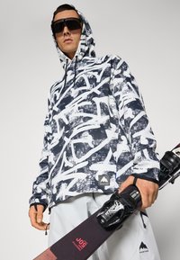 Pullover con cappuccio in un motivo astratto blu e bianco; realizzato in materiale morbido. Presenta un'etichetta con logo di montagna e una mano che tiene attrezzatura per gli sci.