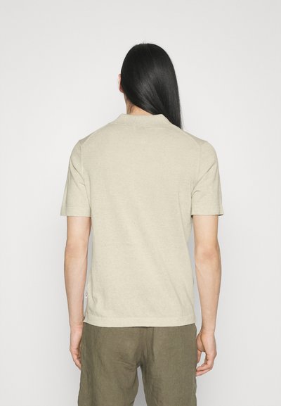 Persona con capelli lunghi e neri che indossa una polo beige a maniche corte e pantaloni verde oliva, di fronte a uno sfondo bianco uniforme.