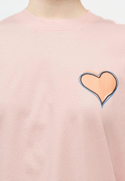Ροζ ανοιχτό μπλουζάκι με ένα στυλιζαρισμένο γραφικό καρδιάς στο αριστερό στήθος, περιγραμμένο με μαύρες, μπλε και πορτοκαλί γραμμές.