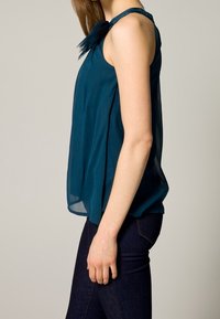 Haut sans manches bleu sarcelle en tissu léger, avec un détail volanté sur l'épaule, au design ample et fluide. Assorti à un jean foncé.