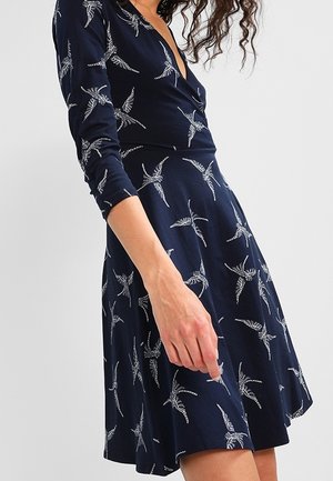 Vrouw draagt een marineblauwe jurk met witte vogelpatronen, driekwart mouwen en een diepe V-hals, getoond van schouder tot halverwege de dij.