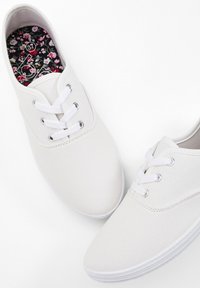 Sneakers bianchi in tela con sei passanti, lacci bianchi e suola in gomma texturizzata. L'interno presenta una soletta con motivo floreale.