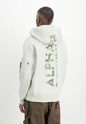 Mann trägt hellgrauen Alpha Industries Hoodie mit großem vertikalem Logo auf dem Rücken und braune Hose, von der Kamera weg gerichtet.