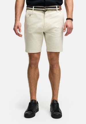 Beige katoenen shorts met een vlakke voorkant, zijzakken en een kleine logo patch; gecombineerd met zwarte lace sneakers en een multi-gestreepte riem.