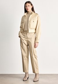 Γυναίκα που στέκεται φορώντας μπεζ κοντό σακάκι, μπεζ ζιβάγκο, μπεζ παντελόνι ψηλής μέσης και μποτάκια ταμπά σε ουδέτερο φόντο.