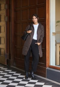 Abrigo largo acolchado marrón con capucha, camiseta blanca, pantalones negros y botines negros. Bolso negro simple, de pie sobre un suelo de cuadros.