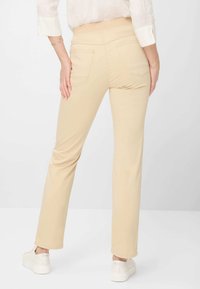Beige stretch pants with a high waistband and two back pockets, paired with white sneakers, displayed from the back.