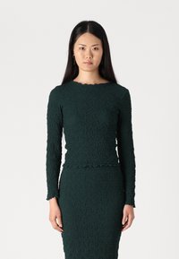 Dark green textured long-sleeve top with a scalloped neckline and hem. Paired with a matching fitted skirt. Smooth fabric with a relaxed fit.
