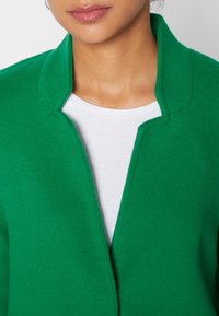 Chaqueta de mezcla de lana verde con una textura suave, que presenta un cuello marcado y un diseño de frente abierto. Llevada sobre una camiseta blanca.
