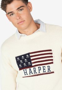 Suéter de punto color crema con un diseño de bandera estadounidense en azul y rojo, con una calavera y tibias blancas, y la palabra "HARPER" debajo.