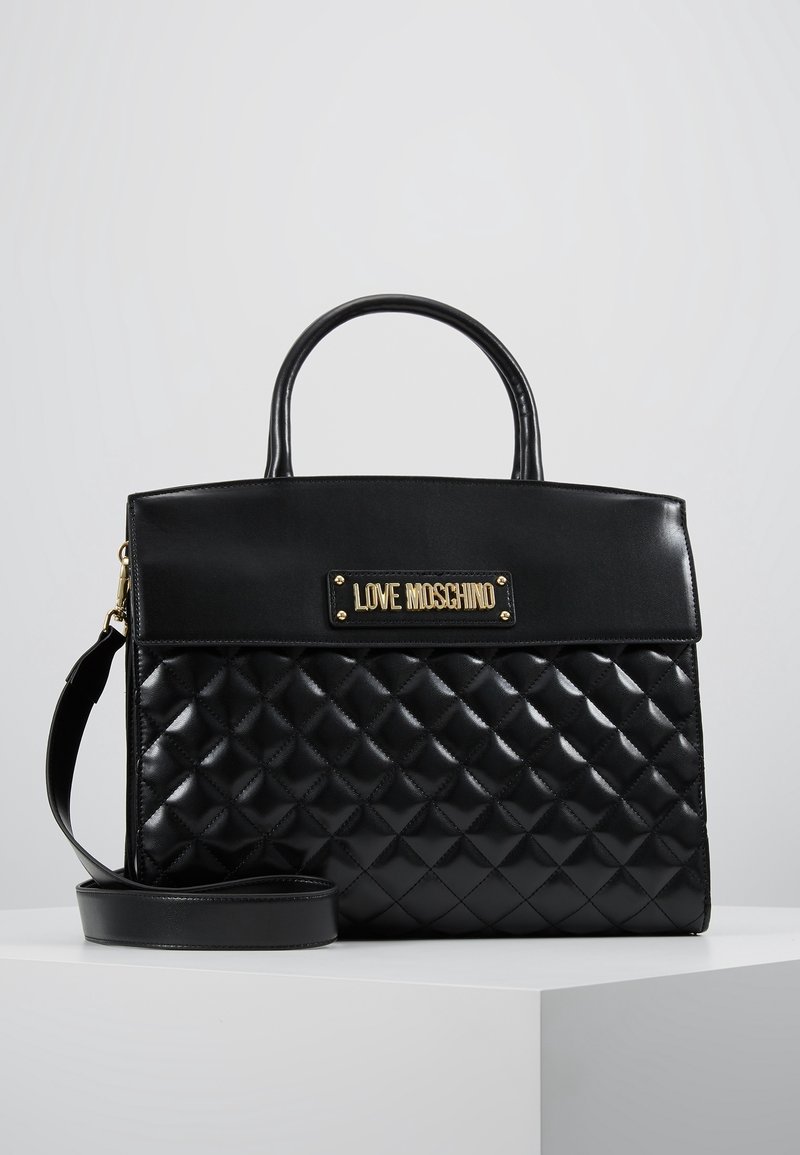 Musta, tilkkutyylinen Love Moschino -käsilaukku, jossa on yläkahvat ja irrotettava olkahihna, esillä valkoisella alustalla.