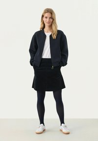 Marineblauw zip-up bomberjack, witte top, zwarte ribfluwelen rok en zwarte panty. Witte sneakers met gumsoles maken de outfit compleet.