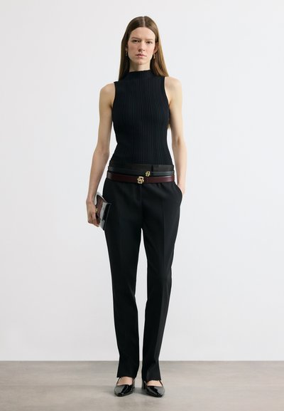 Femme aux longs cheveux raides portant un haut noir sans manches, un pantalon noir avec deux ceintures, tenant un portefeuille marron dans une main.