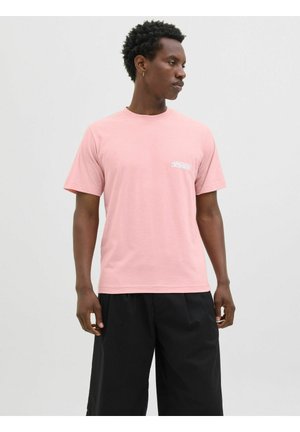 Helles rosa Baumwoll-T-Shirt mit kurzen Ärmeln und Rundhalsausschnitt, mit einem weißen Logo auf der linken Brust, kombiniert mit schwarzen Hosen.
