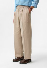 Beige geplooide broeken met een relaxte pasvorm, vervaardigd van een gestructureerde stof. Met zijzakken en gedragen met donkerbruine instappers.