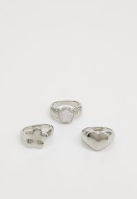 Tres anillos de plata con formas diferentes: un corazón, una flor y un anillo con forma de flor que tiene un centro de piedra iridiscente.