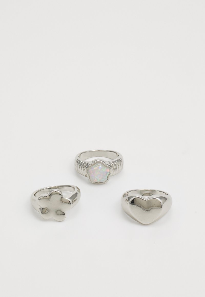 Tres anillos de plata con formas diferentes: un corazón, una flor y un anillo con forma de flor que tiene un centro de piedra iridiscente.