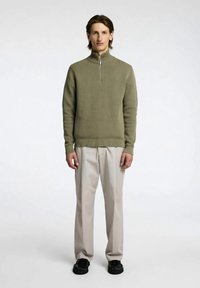 Ζακέτα ελιάς με φερμουάρ και ριμπ υφή, σε συνδυασμό με ανοιχτό μπεζ παντελόνι και μαύρα παπούτσια. Καθαρός, μινιμαλιστικός σχεδιασμός.