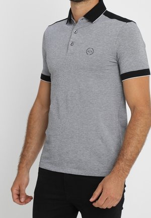 Homme portant un polo gris avec un col et des bordures de manches noirs, orné d'un petit logo "A|X" sur la poitrine.