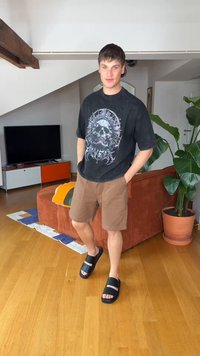 Jeune homme debout à l’intérieur portant un t-shirt noir avec un motif tête de mort, un short marron et des sandales noires, avec une plante et une télévision en arrière-plan.