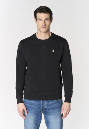 Uomo che indossa una felpa nera a maniche lunghe con piccolo logo bianco sul petto e jeans blu, in piedi davanti a uno sfondo chiaro e uniforme.