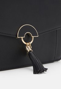 Borsa a tracolla in pelle nera con una chiusura circolare dorata e un accento in pelle a frangia. Texture liscia con dettagli di cucitura minimi.