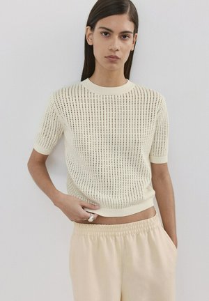 Młoda kobieta ubrana w kremowy sweterkowy top z krótkim rękawem o perforowanym wzorze oraz beżowe spodnie z elastycznym pasem, stojąca na tle jednolitej ściany.