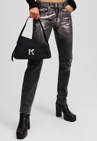 Sac à main en denim noir avec quincaillerie argentée, de forme triangulaire, et avec un logo "K" bien en vue. Modèle portant un jeans gris métallique et des bottes à plateforme noires.
