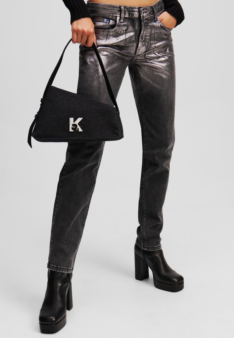 Sac à main en denim noir avec quincaillerie argentée, de forme triangulaire, et avec un logo "K" bien en vue. Modèle portant un jeans gris métallique et des bottes à plateforme noires.