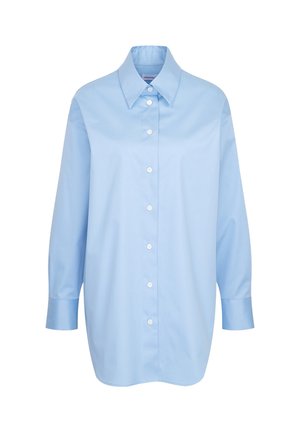 Personne aux cheveux courts et bouclés portant une chemise ample bleu clair boutonnée, vue de dos sur un fond beige.
