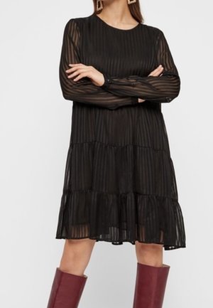 Mujer vestida con un vestido negro translúcido de rayas, manga larga y falda en capas, combinada con botas burdeos hasta la rodilla, brazos cruzados.