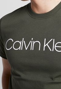 Olive zelené bavlněné tričko s velkým bílým nápisem "Calvin Klein", kulatým límcem a krátkými rukávy s hladkou texturou.