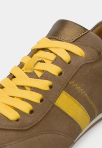 Καφέ σουέντ αθλητικό παπούτσι με κίτρινα κορδόνια και πλαϊνή ρίγα, που διαθέτει μεταλλική λεπτομέρεια στη φτέρνα και επιφάνεια με υφή.